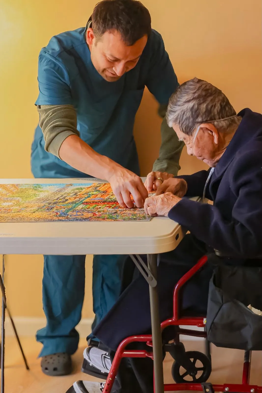 Two people working together on a puzzle at a table, with a wheelchair visible beside it